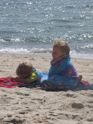 Zion lying on the beach, Lijah sitting wrapped in a  towel