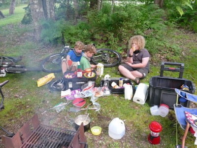 the boys sitting on the ground among all the STUFF at the campsite, having dinner
