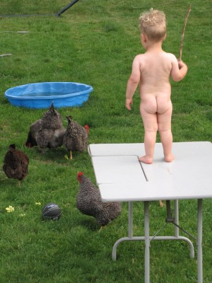 Naked Lijah standing on top of a table on the lawn, holding a stick, watching the chickens