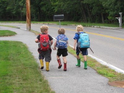 Harvey and two friends walking down the sidewalk in backpacks and rain boots