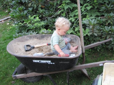 Lijah playing in a dirt-filled wheelbarrow, with shovels