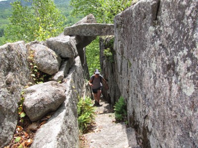 Becca climbing up an exposed path, passing under a block of granite