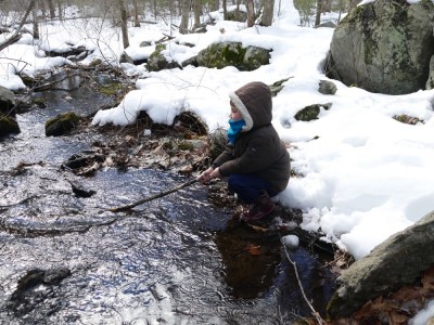 Lijah on the snowy bank holding a stick over a brook