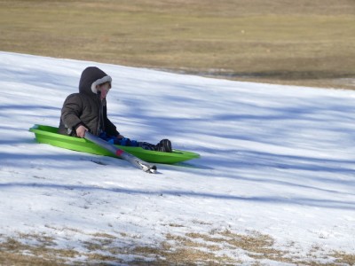 Lijah sledding down a narrow band of snow on a grassy hill