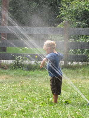 Zion, in his clothes, standing with his back to the sprinkler stream getting soaked
