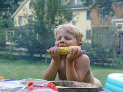 Zion at the picnic table taking a bite of corn on the cob with his eyes closed