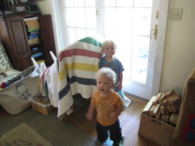 Zion and Elijah standing outside their house made from a blanket draped over the tipped-over rocking chair