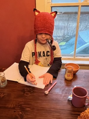 Elijah writing at the kitchen table wearing a fox hat with a pipe in his mouth