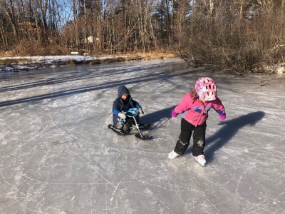 Lijah sitting on a sled on the ice getting pulled by a friend on skates