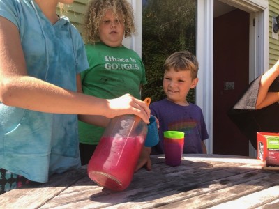 Harvey and Elijah watching a friend pour from a jar of purple juice