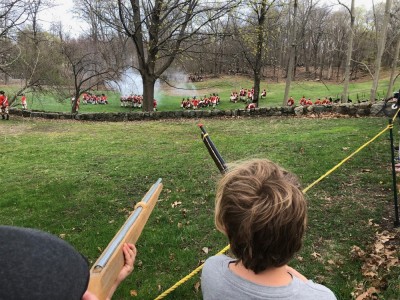 Zion and Elijah aiming muskets at a group of Redcoats fighting a battle