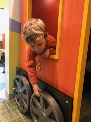 Lijah, in his orange sweater, leaning out the window of the orange play train at the museum