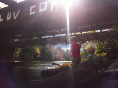Harvey and Zion playing by the river, with the sun, fall trees, and a rail bridge in the background