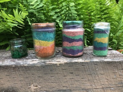 jars filled with stripes of colored sand sitting in front of ferns