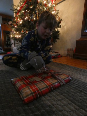 Elijah wearing shark mittens opening a present in front of the tree