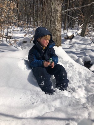 Elijah sitting in the snow in the woods eating a granola bar