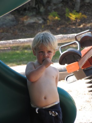 Zion, shirt off, sucks his thumb standing on the playground